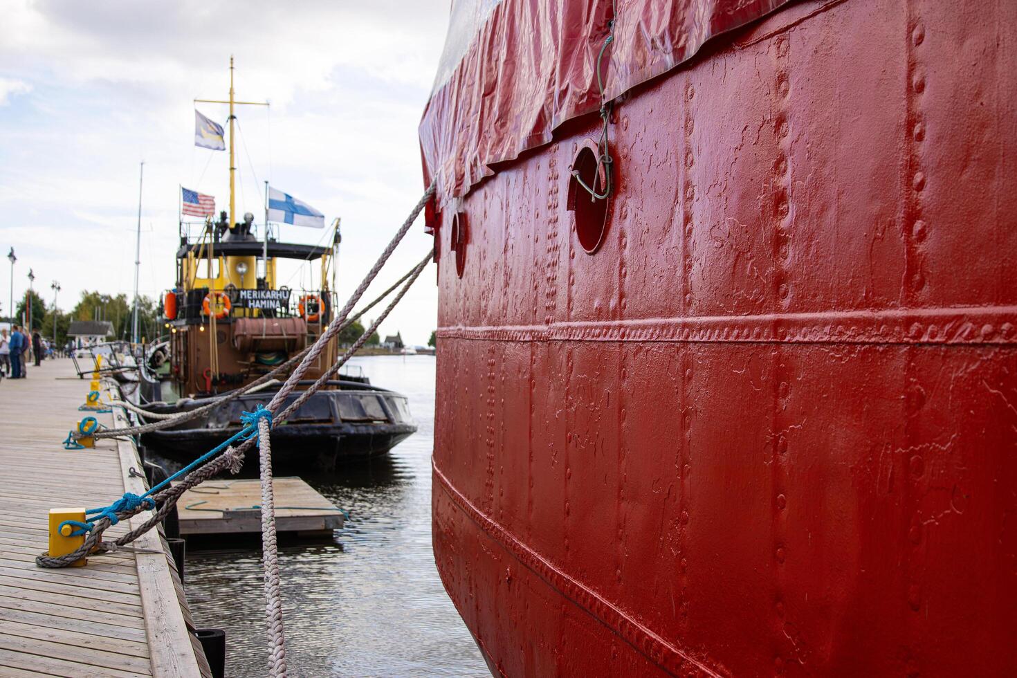 Tervasaari, Hamina, Finland, August 26th, 2023. The red ship docked at wooden pier, tied securely with thick marine ropes. The famous vessel Merikarhu on the background photo