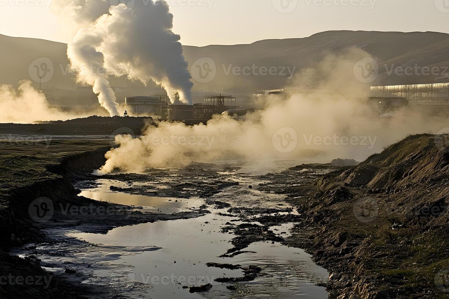 Industrial landscape showing steam and smoke rising from pipes, with a river and surrounding hills in the background during golden hour lighting conditions photo