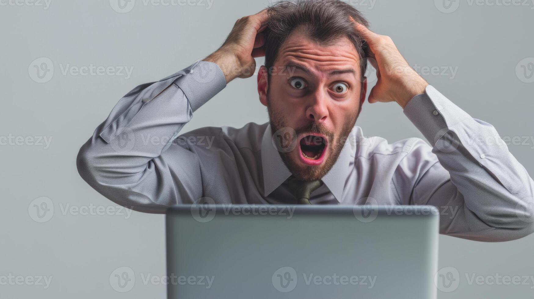 man in a shirt and tie is sitting at a desk with a laptop, hands on his head, looking shocked or stressed with his mouth wide open. photo