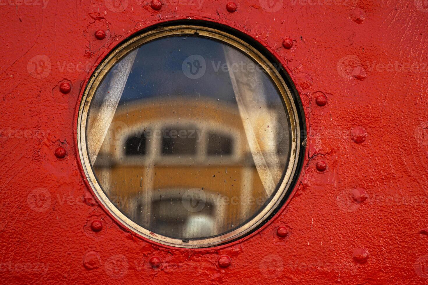 A detailed closeup of a circular ship porthole set in a vivid red metal wall, reflecting the yellow and white facade of the historic customs office in Tervasaari harbor, Hamina, Finland. 17 photo