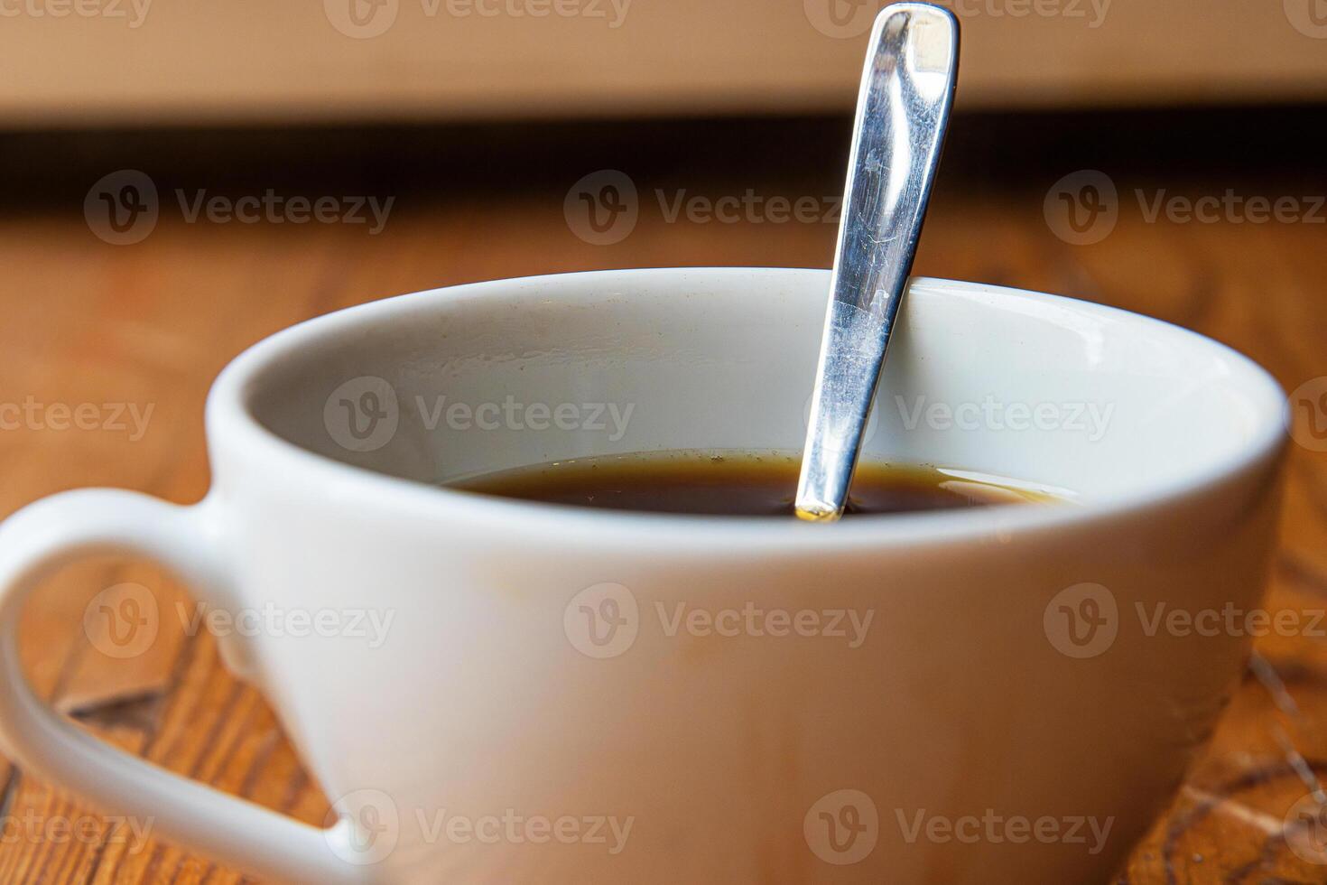 A closeup of a white ceramic cup filled with black coffee, with a metal spoon inside, placed on a rustic wooden table.Soft focus 10 photo