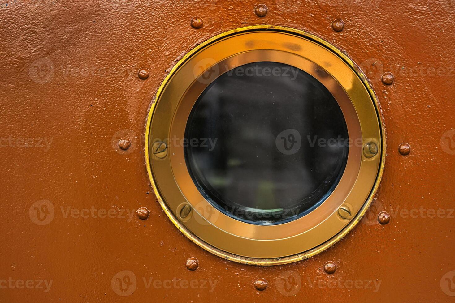 Round brass framed porthole window embedded in a textured rust colored ship wall. Contrast between polished metal and aged surface photo