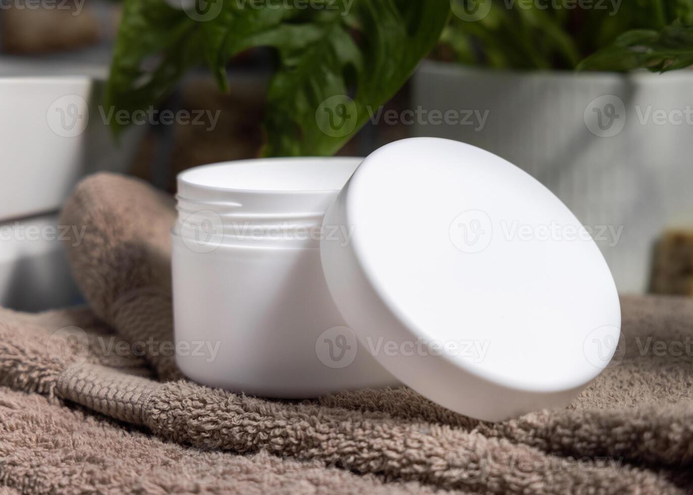 Opened cream jar with blank lid on brown bath towel near green plant, cosmetic mockup photo