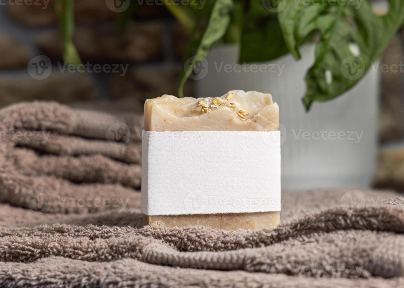 Soap with blank label on brown folded towels near green plant in bathroom, mockup photo