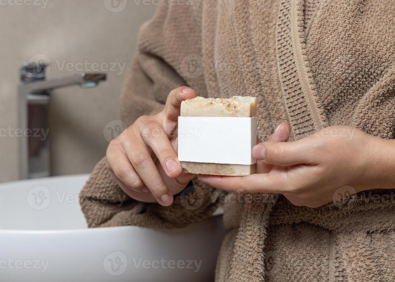 Closeup of woman in brown bathrobe with soap with a label in hands, mockup photo