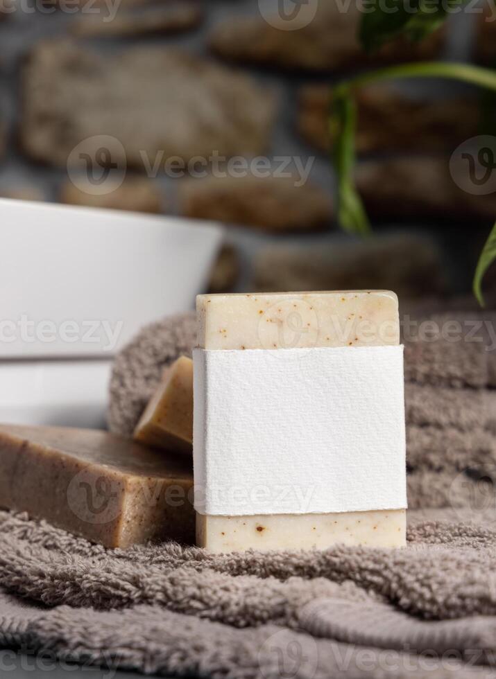 Soap with a label on brown folded towels near plant and basin in bathroom, mockup photo