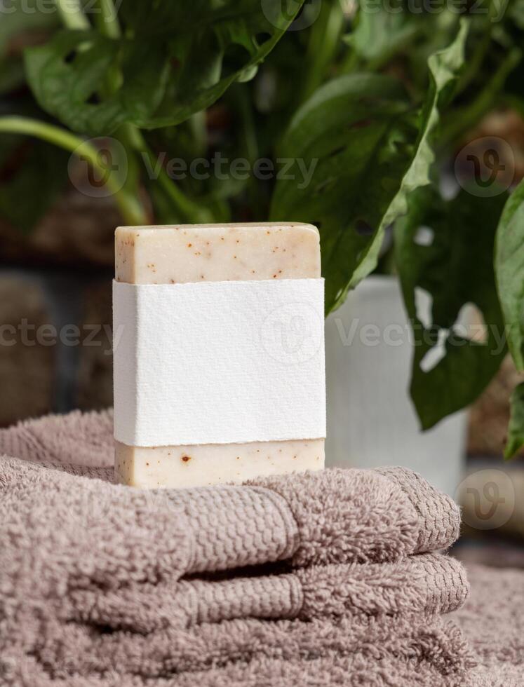 Soap bar with a label on brown folded towels near plant in bathroom, mockup photo