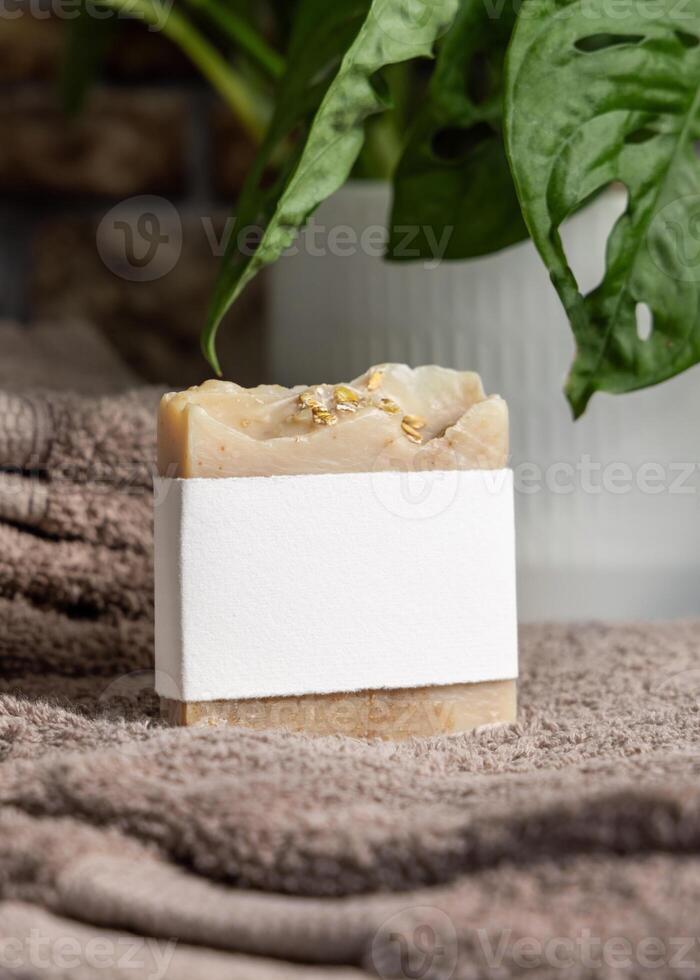 Soap bar with blank label on brown towels near green plant in bathroom, mockup photo