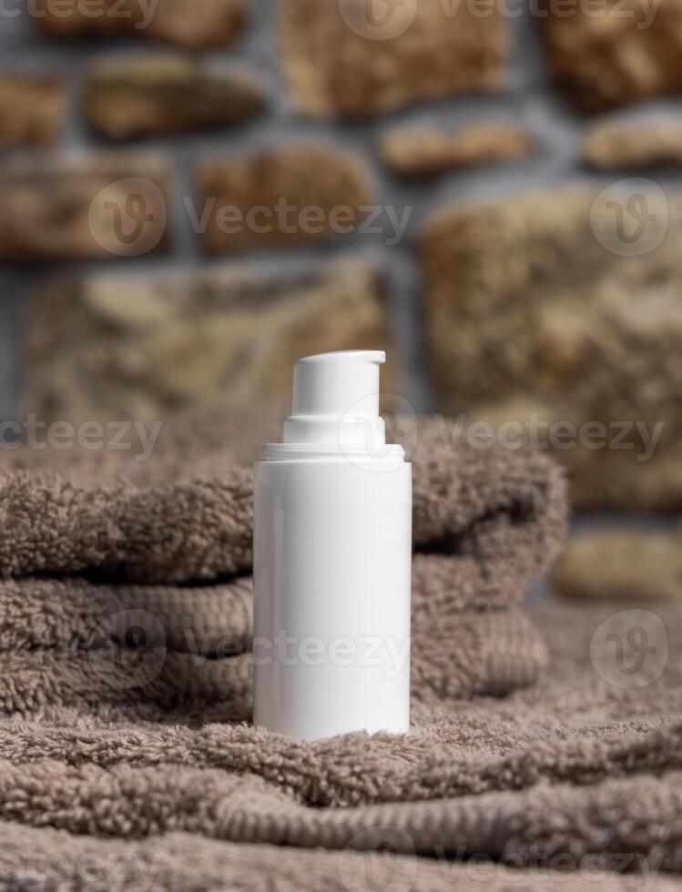 White cream pump bottle on brown folded towel against stone wall close up, cosmetic mockup photo