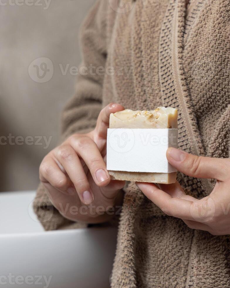 Closeup of woman in light brown bathrobe with soap bar with blank label in hands, mockup photo