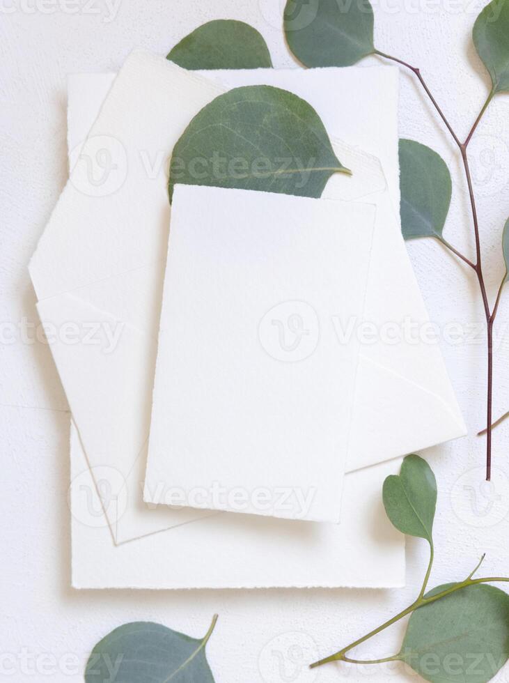Card and envelope on a white table near green eucalyptus branches and leaves top view, Wedding mockup photo