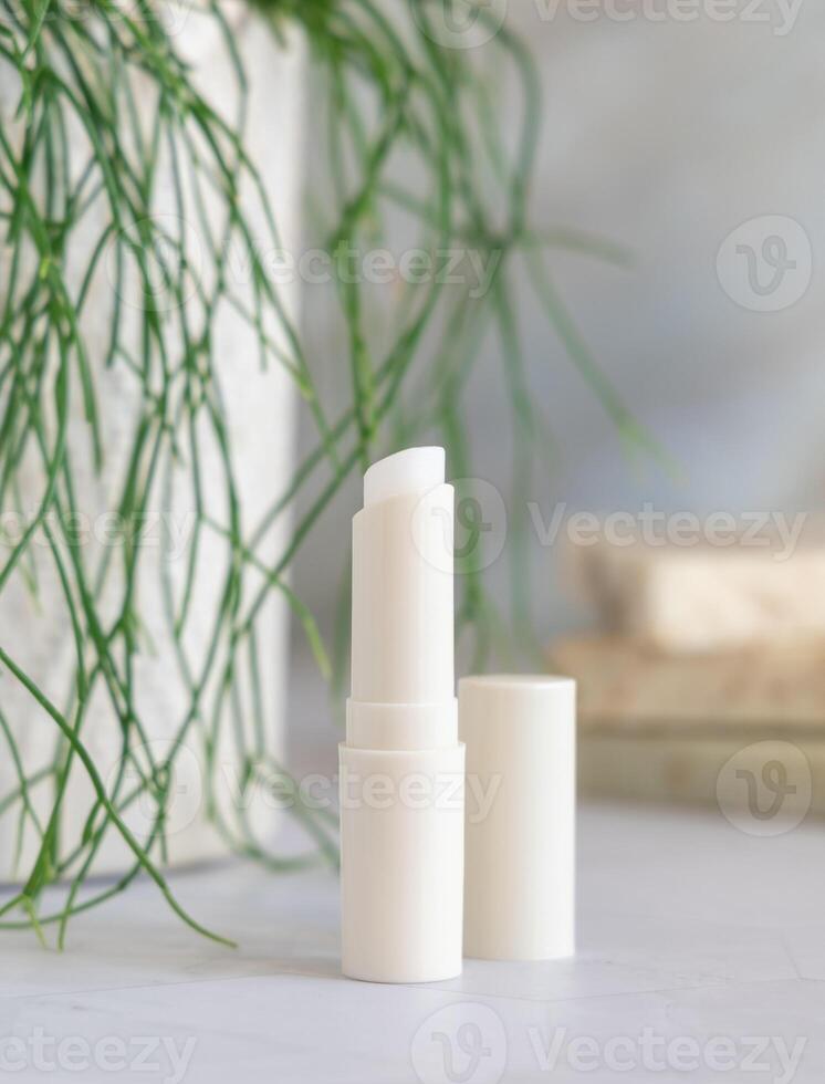Opened lip balm stick on marble near green plant in bathroom, mockup photo