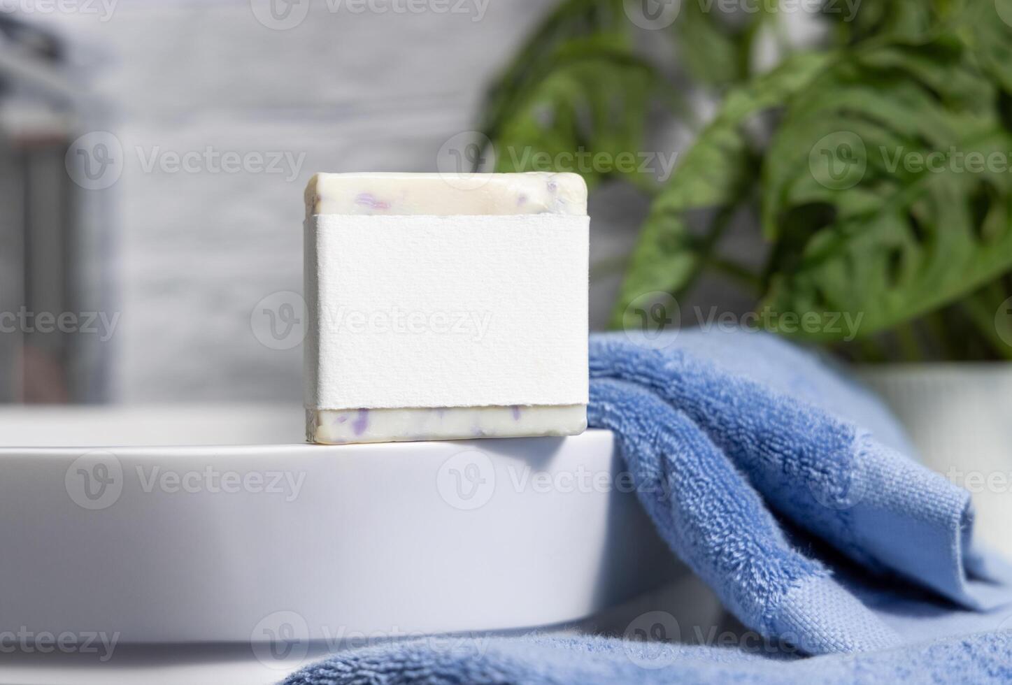 Soap bar with blank label on sink near blue towels and green plant close up, mockup photo