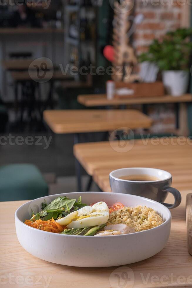Bowl of couscous, meat, eggs and avocado and vegetables and cup of coffee on cafe table photo