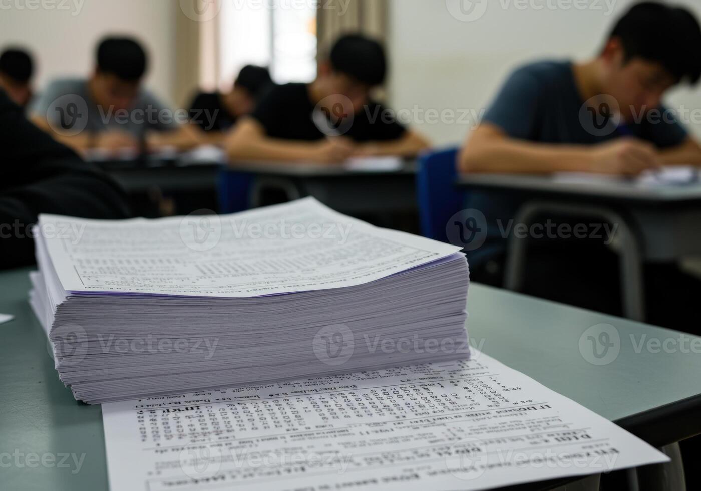 A stack of exam papers sits on a desk as students take a test in the classroom setting. photo