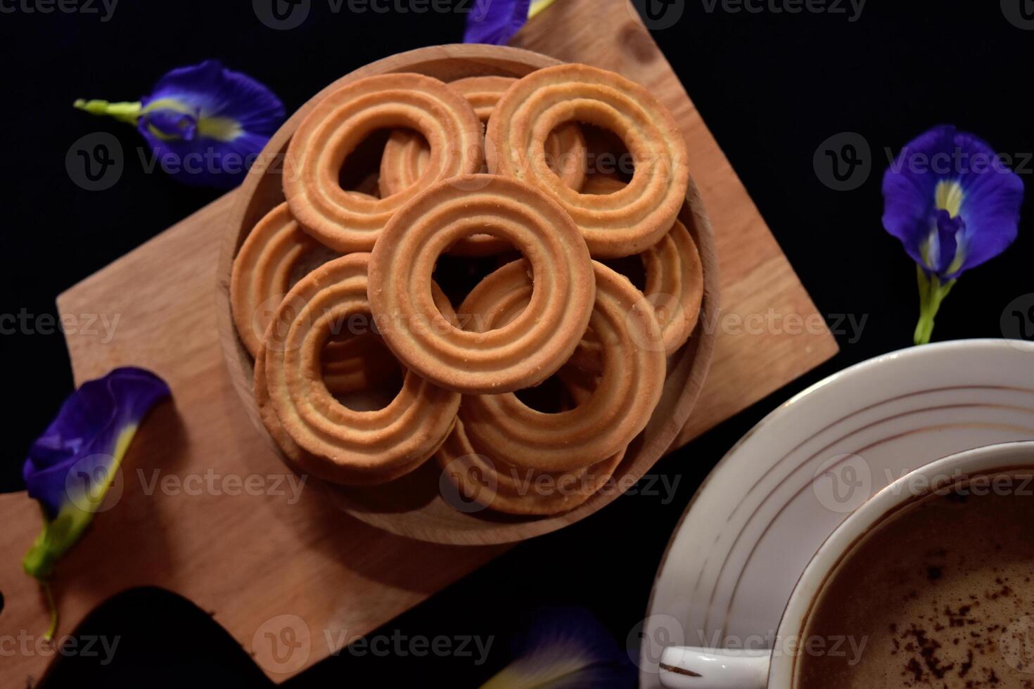 Closeup view of a bunch of ring biscuits on a wooden tray with some butterfly pea flowers as decoration. Dark mood concept of food photography. photo