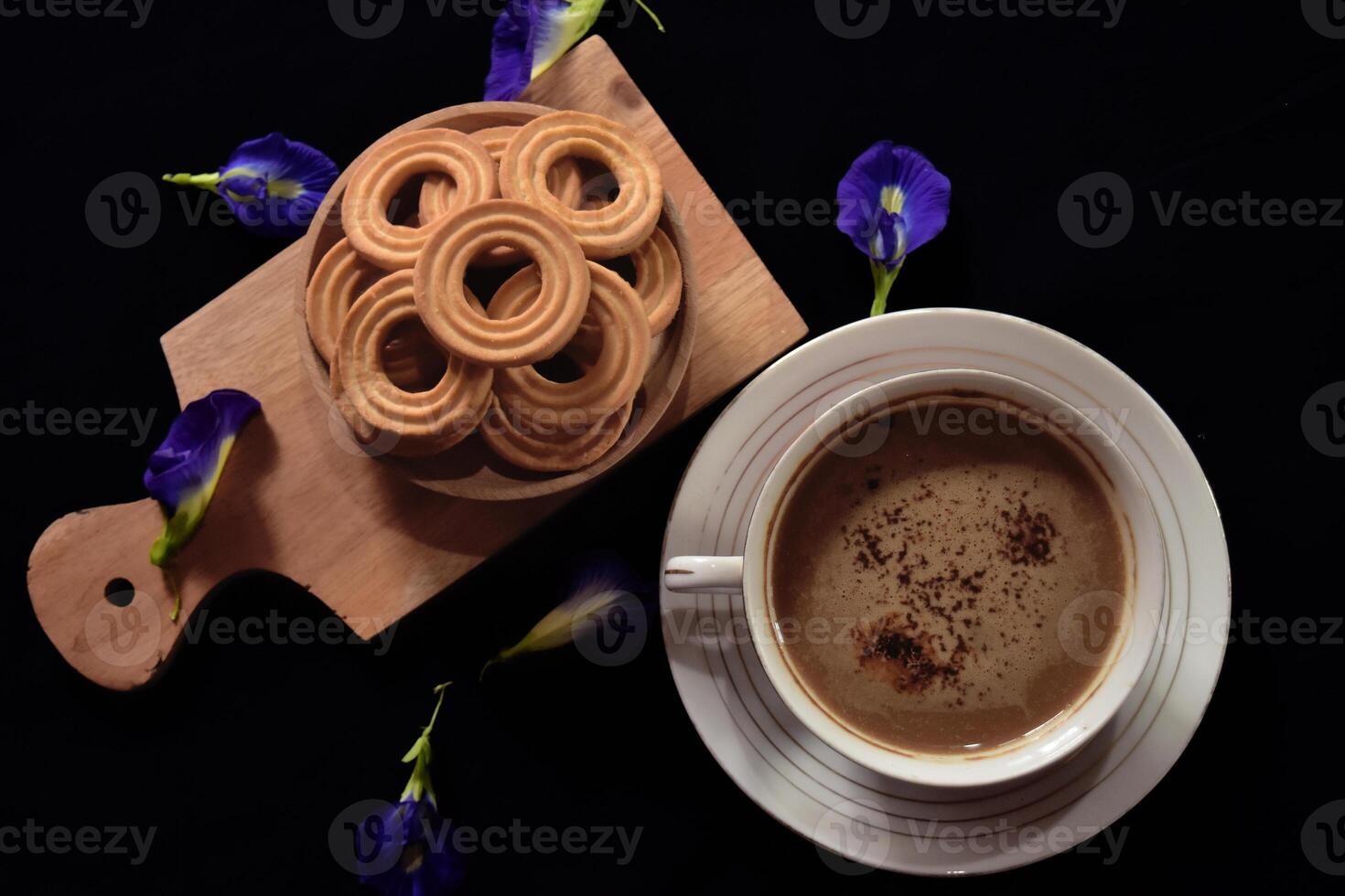 Flat lay view of a bunch of butter cookies on a wooden tray and a cup of cappuccino with some blue pea flowers as decoration. Dark mood concept of food photography. photo