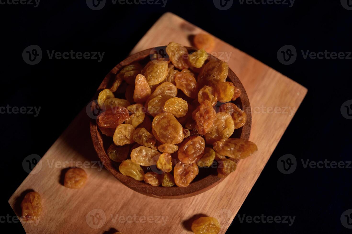 A bowl of raisins on a wooden cutting board with a dark background. Food photography of a bunch of dried grapes commonly used in cooking and baking. photo
