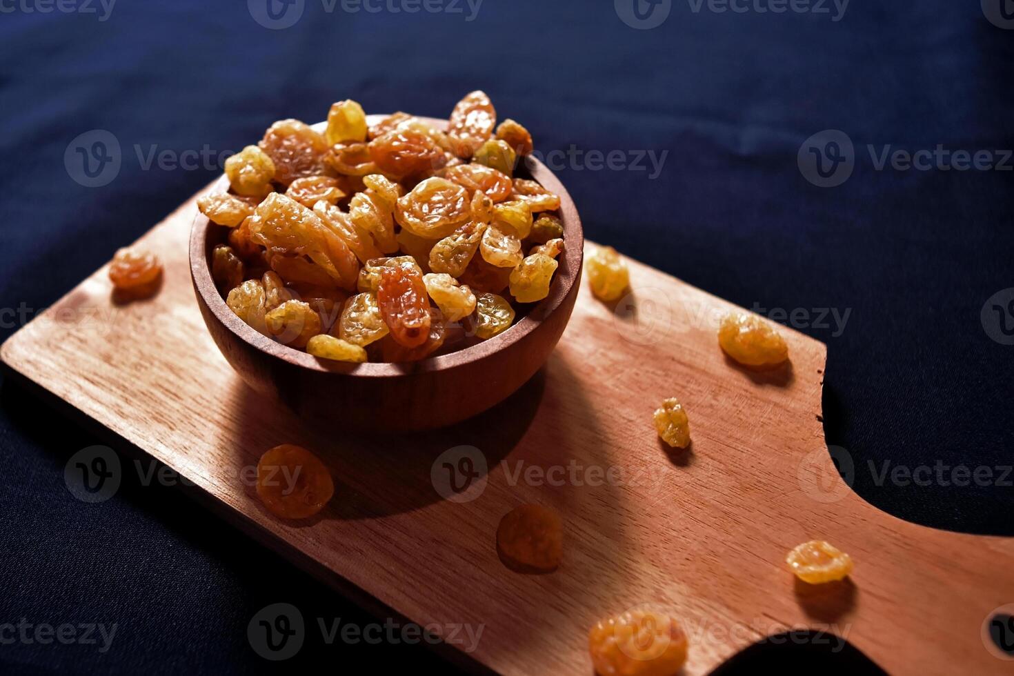 Closeup view of a bowl of raisins on a wooden cutting board with a dark background. Food photography of a bunch of dried grapes commonly used in cooking and baking. photo