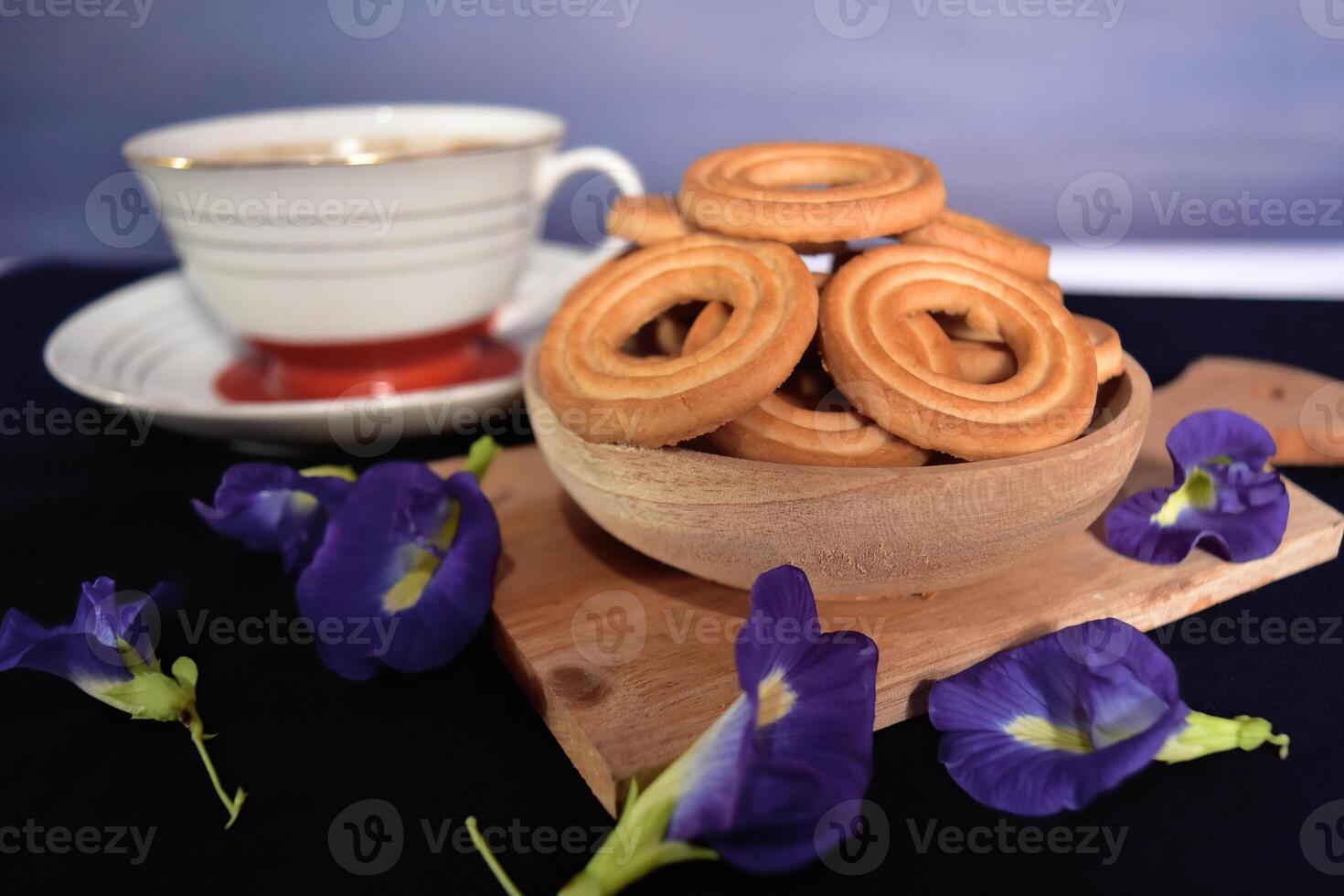 A bunch of butter cookies on a wooden tray and a cup of coffee with some butterfly pea flowers as decoration. Dark mood concept of food photography. photo