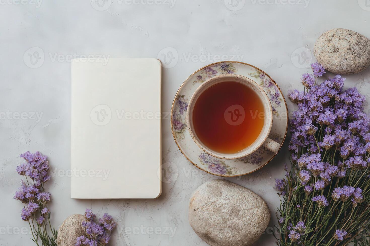 A serene setting features a cup of tea beside a blank notebook and sprigs of purple flowers. Smooth stones surround the arrangement, evoking a calm and creative moment. photo