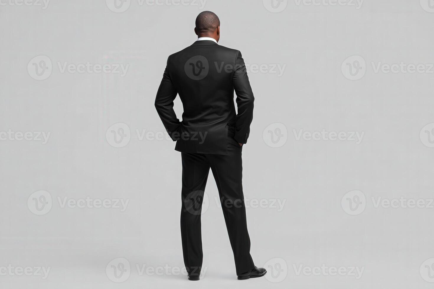 A man wearing a sleek black suit stands with his back to the viewer, exuding confidence and professionalism. The backdrop is minimalist, enhancing the focus on his attire and posture. photo