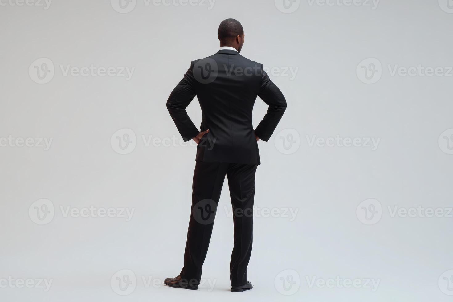 A man wearing a formal black suit stands with his back to the viewer, hands resting on hips. His posture conveys confidence. The setting features a simple and neutral background. photo