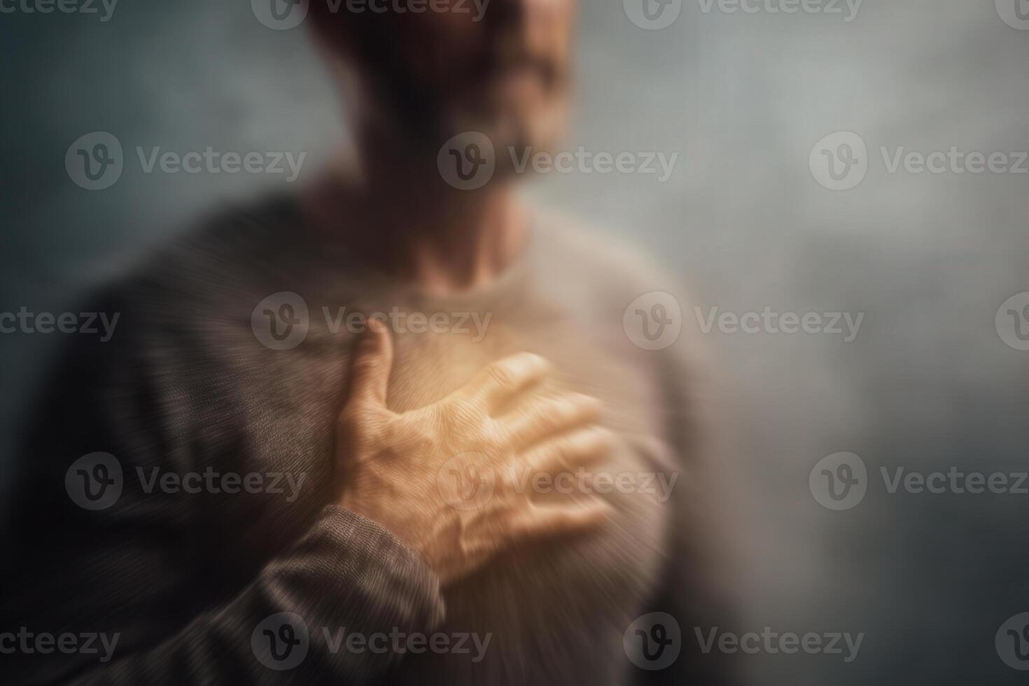 A man stands in a soft mist, placing his hand on his chest as he seems to engage in introspective thought. The muted tones and blurred background create a sense of calm and reflection. photo