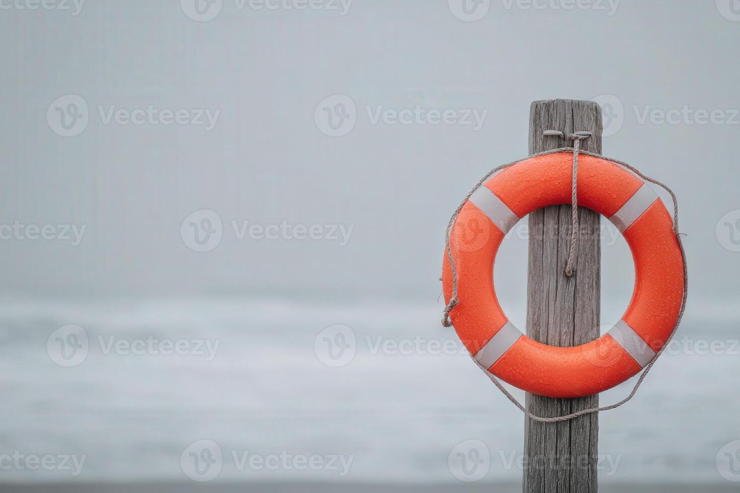 A bright orange lifebuoy hangs on a weathered wooden post at the edge of a tranquil beach. The ocean waves rest gently in the background, under a cloudy sky, creating a serene atmosphere. photo