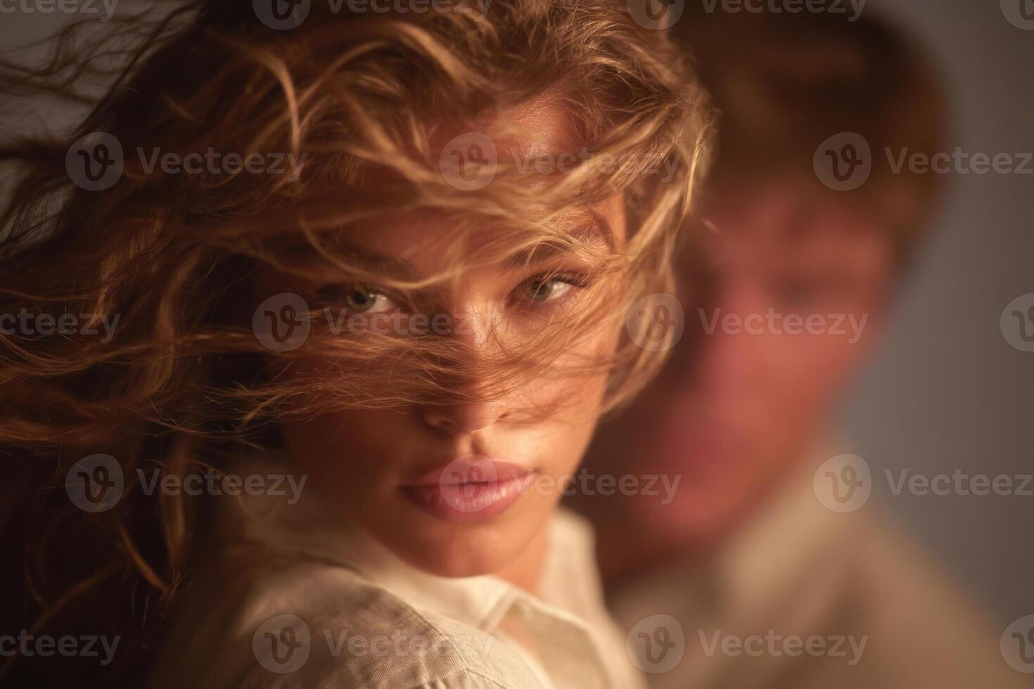 A woman with long, wavy hair stands outside, her face illuminated by soft light, as her hair is caught by the wind. A blurred figure appears in the background, creating a sense of depth and intimacy. photo