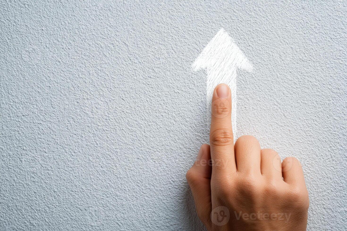 A hand with a finger extended points towards a white arrow sketch on a gray wall. The gesture suggests direction or an upward movement, creating an engaging visual focus. photo