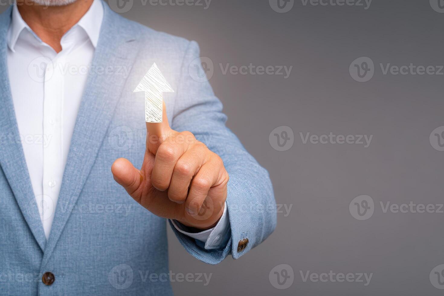 A man in a light blue suit points towards a digital upward arrow symbol, representing growth or advancement in a professional context. The backdrop is a simple gray hue. photo