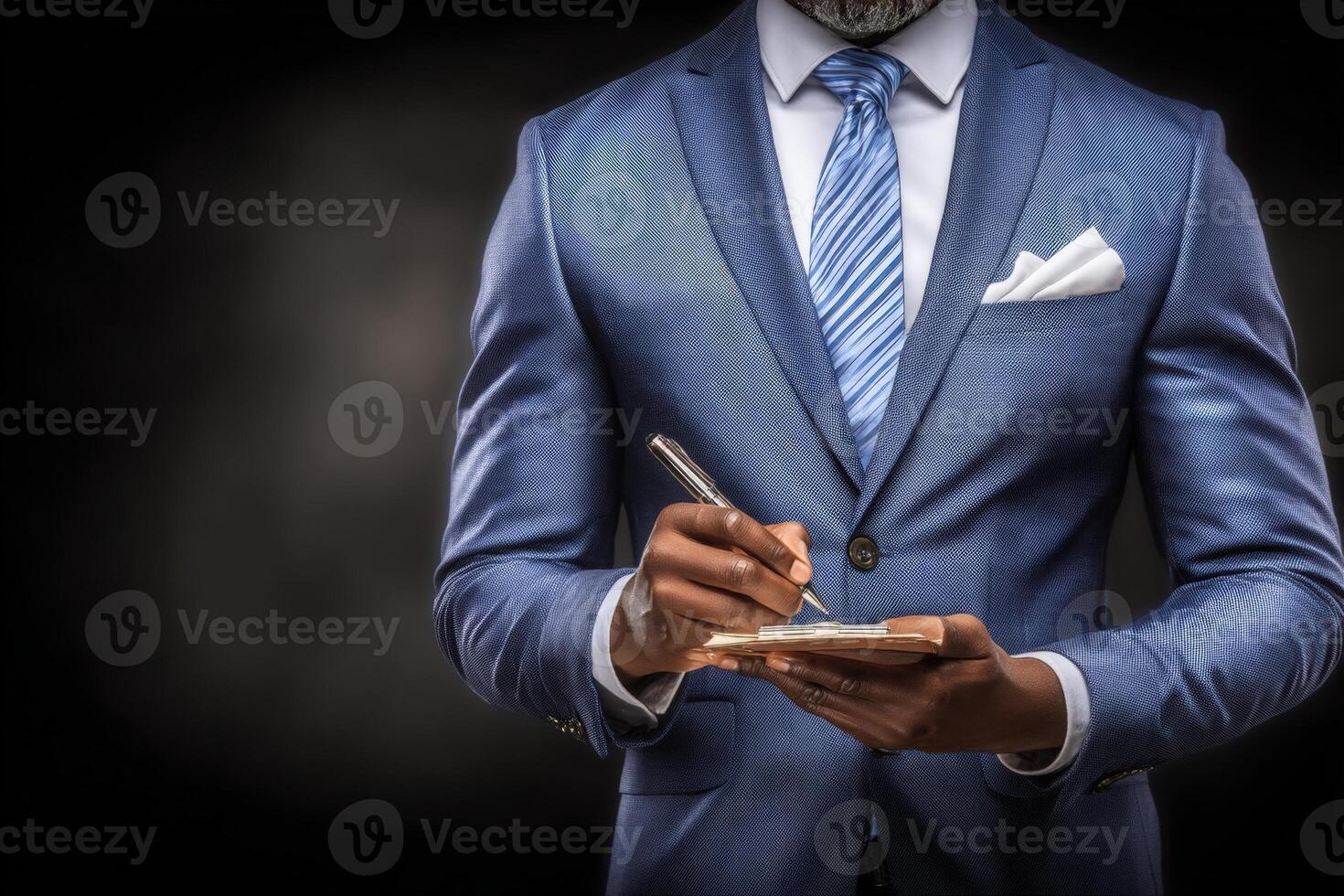 A man in a tailored blue suit stands against a dim background, writing in a notepad with a pen. His stylish appearance is complemented by a blue tie and a white pocket square. photo