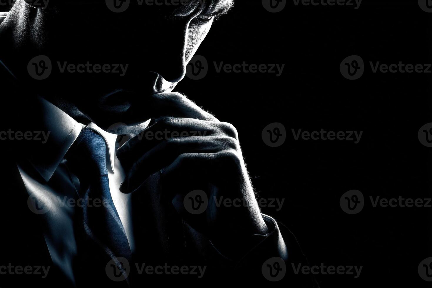 A man dressed in a suit appears deep in thought, with his head bowed and hand resting on his chin. The strong contrast of light and shadow creates a dramatic atmosphere, emphasizing his introspection. photo