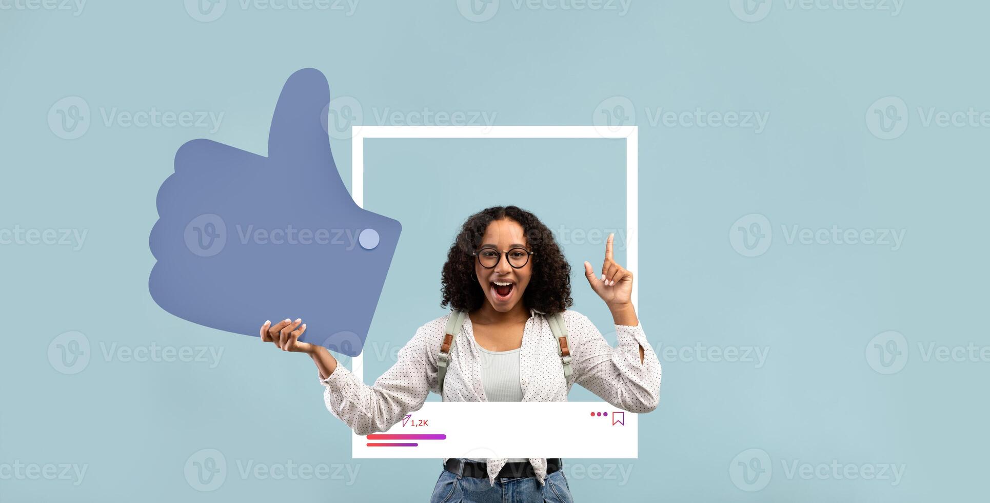 A young woman with curly hair and glasses expresses excitement while holding a large thumbs up symbol in front of a light blue backdrop. She is wearing a casual outfit and has a joyful attitude. photo