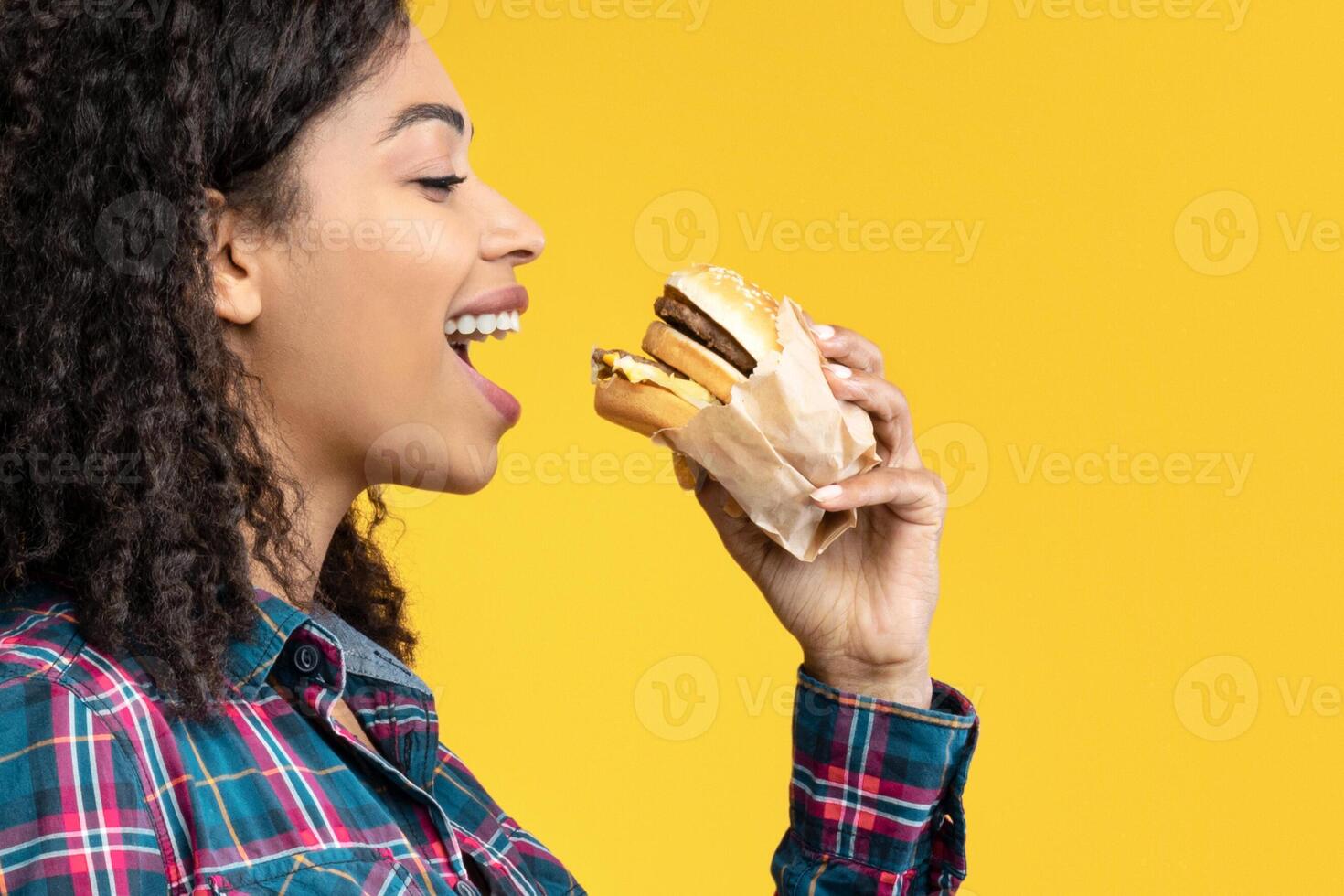 Time For Snack. Side View Portrait Of Hungry Black Lady Eating Hamburger. African American Woman Taking Bite Of Tasty Fast Food, Isolated Over Orange Studio Background, Banner, Copy Space photo