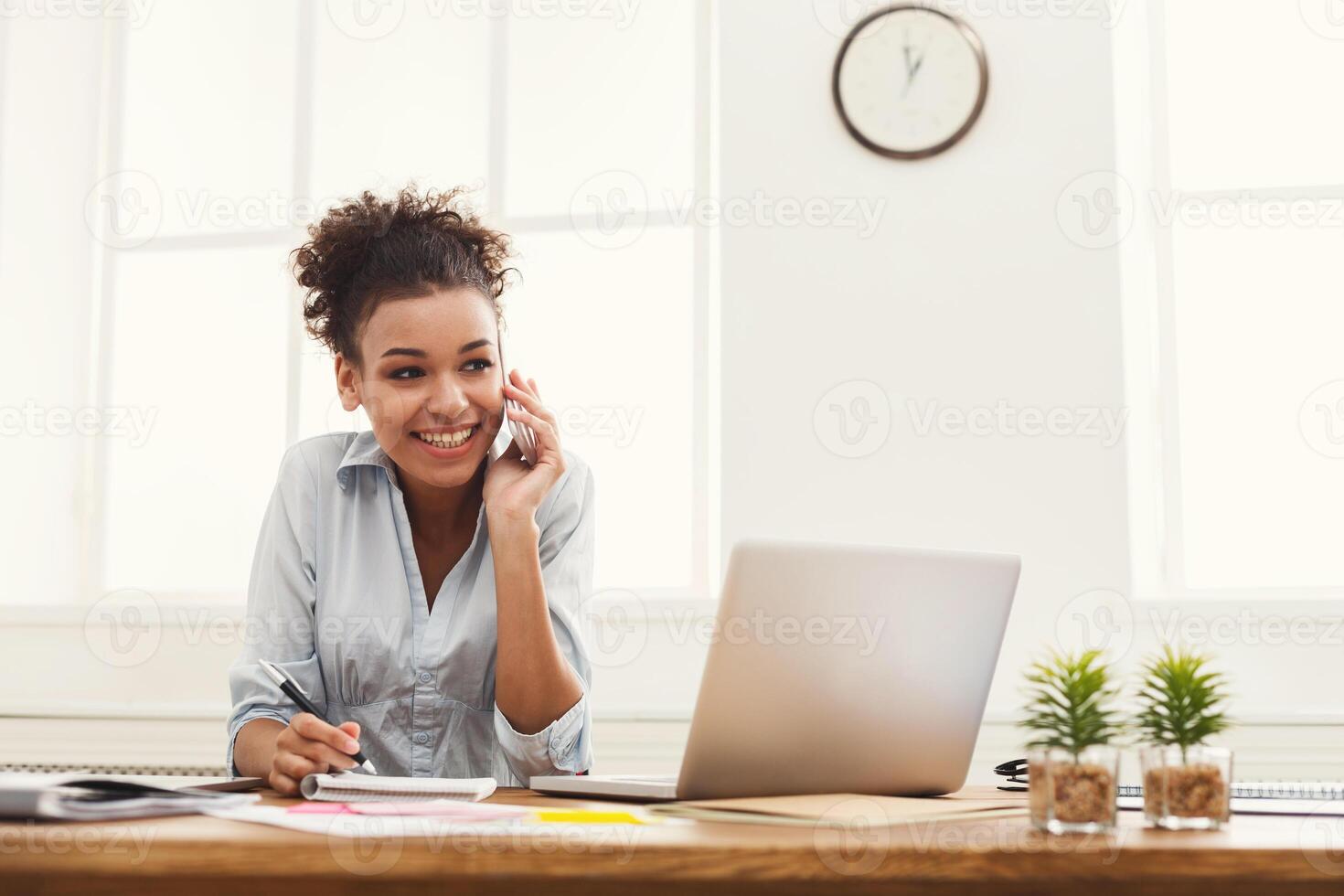 Happy smiling african-american business woman at work talking on phone and taking notes, sitting at her working place in office, copy space photo