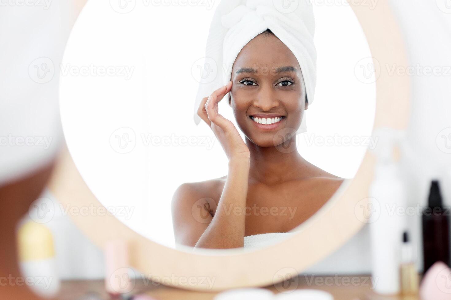 Procedures for beauty, youth and health. Reflection in mirror of fresh young african american lady with towel, smears face with cream after shower, in bedroom in morning at table with cosmetics photo