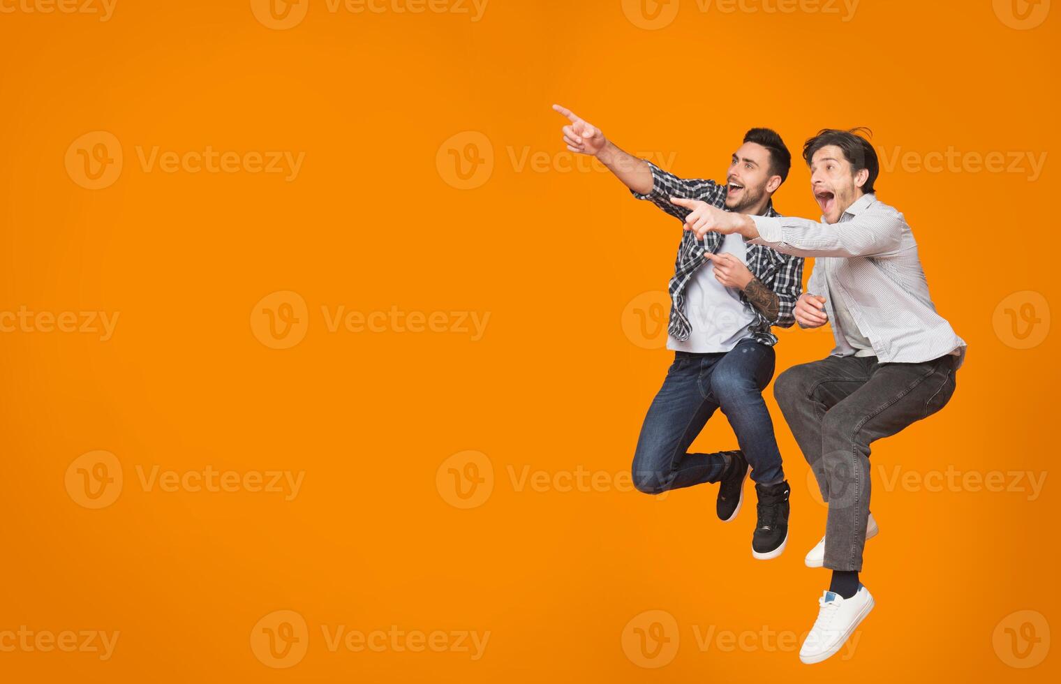 Overjoyed guys pointing at free space and jumping over orange studio background photo
