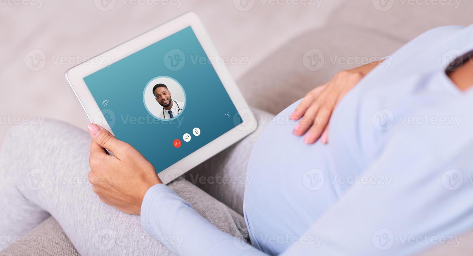 Back view close up of pregnant woman having online consultation at home with black man doctor using tablet photo