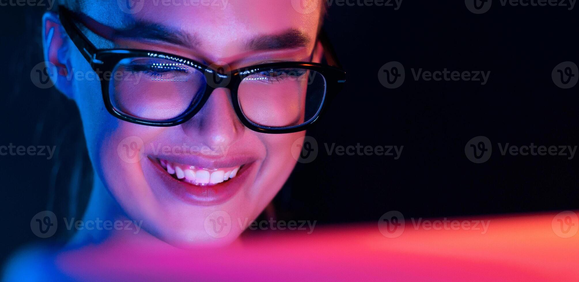 Happy girl looking at laptop screen at night, wearing glasses, face illuminated by neon light, free space photo