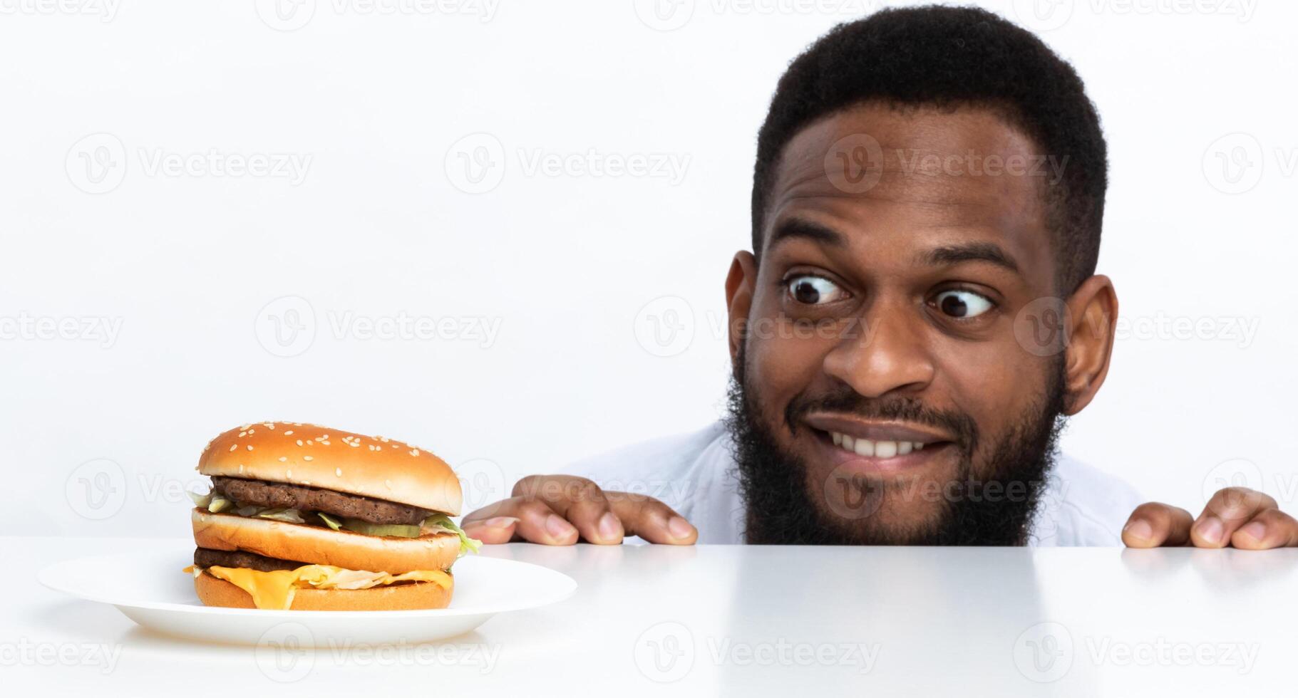 Funny Hungry Black Man Looking At Burger On Table Posing Over White Studio Background. Nutrition And Dieting, Cheat Meal And Unhealthy Junk Food, Overeating Habit Concept. Panorama photo