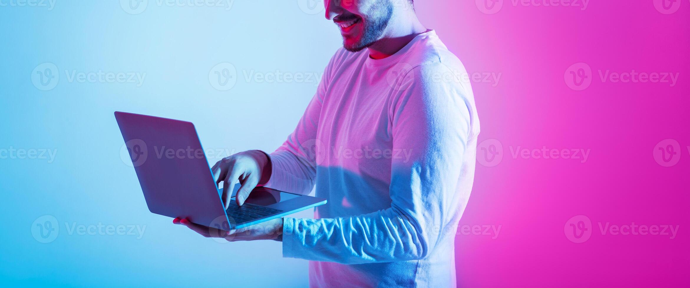 Freelance and work remotely, online game and correspondence. Contented millennial guy use laptop and holds in hands on pink and blue background, panorama, copy space photo
