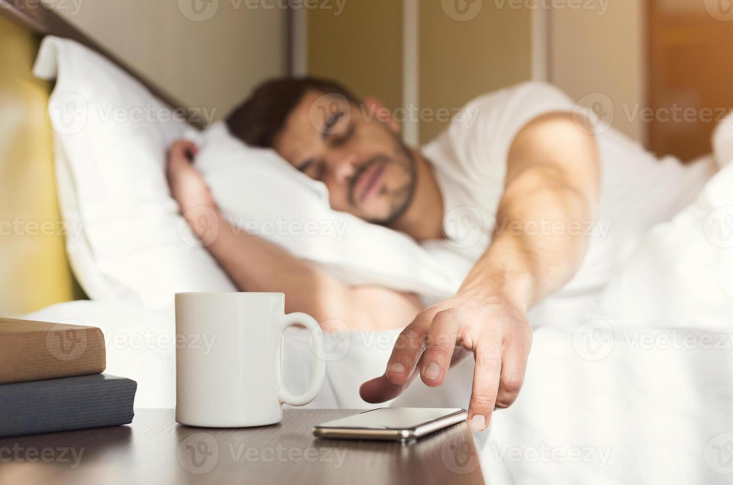 Sleepy guy waking up early after hearing alarm clock signal on smartphone on monday morning, reaching for ringing mobile phone with closed eyes, copy space photo