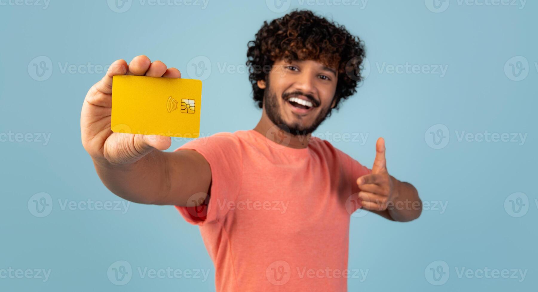 Cheerful handsome young indian man holding yellow credit card and showing thumb up, pleased client demonstrating bank product over blue studio background, panorama. Finance, loan, savings concept photo