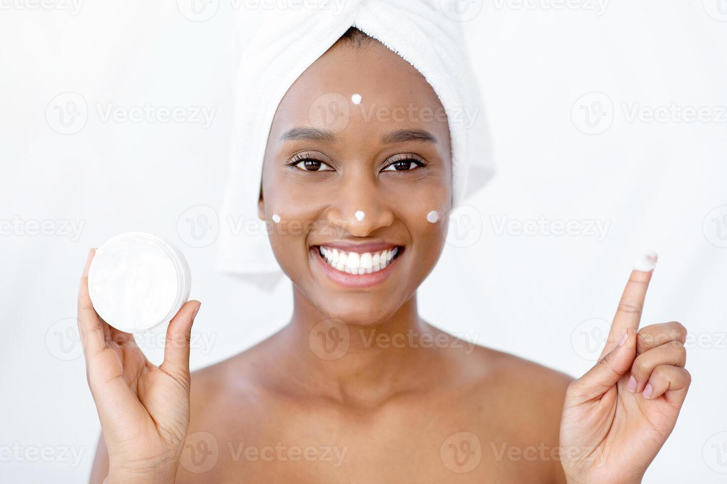 Beauty lessons and facial skin care at home during covid-19 quarantine. Millennial smiling african american lady blogger showing moisturizer cream for face in morning at home and looking in mirror photo