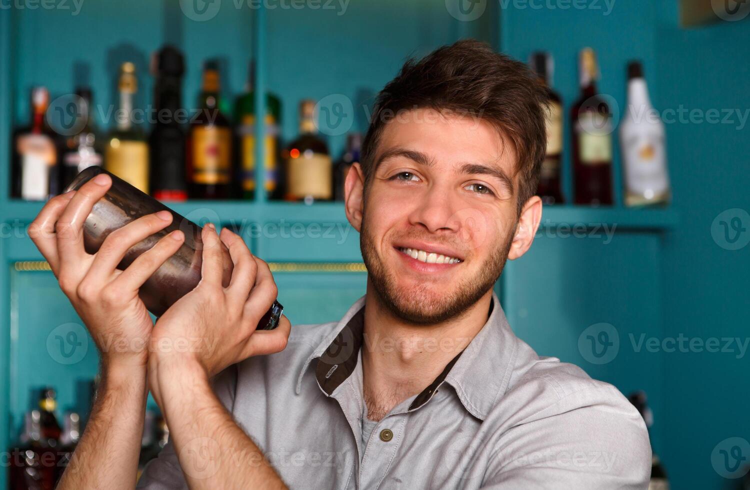 Young handsome smiling barman in bar interior shaking and mixing alcohol cocktail. Professional bartender portrait at work in night club with shaker in hands. Service industry occupation photo