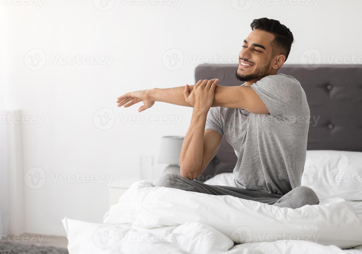 Happy Young Middle Eastern Guy Waking Up In The Morning And Stretching In Bed, Handsome Millennial Arab Man Resting In Bedroom In The Morning After Good Sleep, Ready For New Day, Copy Space photo
