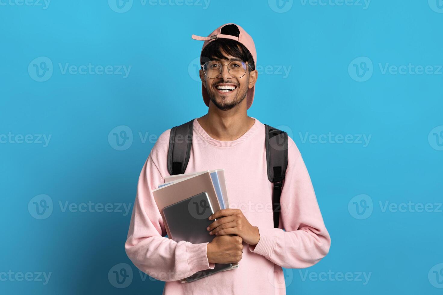 Positive millennial indian guy student in pink baseball cap and sweatshirt posing on blue background, holding books and notepads, carrying backpack, copy space. Education, school, college concept photo