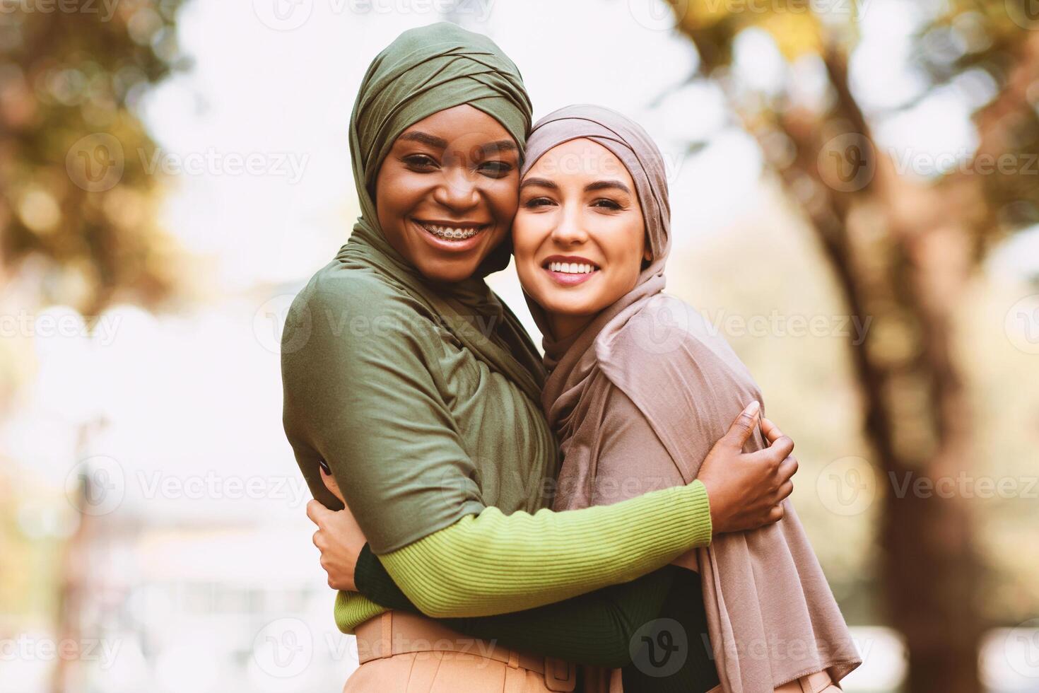 Two Muslim Ladies In Hijab Hugging Smiling To Camera Posing Standing In Park Outdoors. Female Friendship And Togetherness Concept. Black And White Islamic Women Embracing. Panorama, Copy Space photo